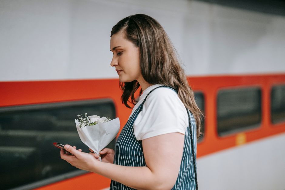 A young woman with long, dark hair is holding a small bouquet of fresh white flowers wrapped in white paper, while standing at a train station platform. She is wearing a white t-shirt layered under a 