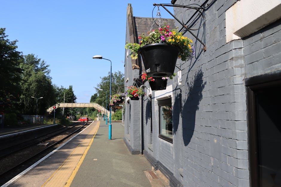 A close-up view of hanging black flower baskets filled with vibrant pink, yellow, and purple blossoms, mounted on the grey brick exterior of a building at Norbiton Station. The arrangements feature a 