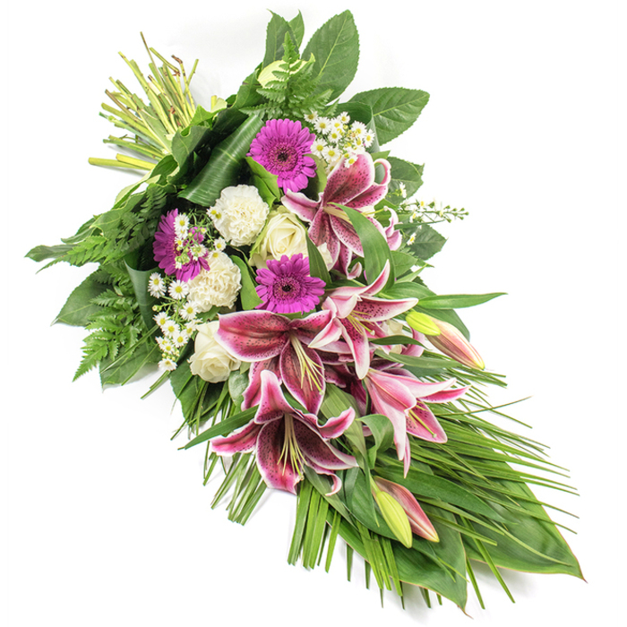 Set against a bright white background, this substantial funeral spray creates a striking diagonal silhouette, flowing from the bound stems at the upper left to the tapering foliage at the lower right. The composition is dominated by large, star-shaped Stargazer lilies whose petals are a vivid fuchsia-pink, their centres heavily speckled with burgundy, then fading to delicate white edges. Some blooms are fully open, offering a sense of openness and heartfelt tribute, while others remain as elongated buds tinged with pink and pale green, promising beauty yet to unfold. Intermingled with these lilies are clusters of magenta gerbera daisies, their flat, cheerful faces bringing a gentle brightness to an arrangement intended for moments of sorrow. White roses, both in tight buds and partially open forms, lend a quiet elegance, while ruffled white carnations add fullness and a soft, cushiony texture that cradles the more striking flowers. Scattered throughout are tiny white filler flowers, reminiscent of daisies or gypsophila, which sprinkle light and delicacy between the larger shapes. The greenery is abundant and carefully structured: broad, dark green leaves create a strong backdrop, fern fronds trail softly along the edges, and long, slender blades of foliage arc outward, emphasising the elongated, teardrop-like form. The visible tying point at the top left shows the stems neatly gathered, reinforcing the hand-tied, artisanal nature of the design. This arrangement feels calm and reverent, ideal for farewells at local chapels or cemeteries around Norbiton, from quiet corners near Kingston Road to the green spaces leading towards Richmond Park, offering a dignified floral expression of sympathy and remembrance.